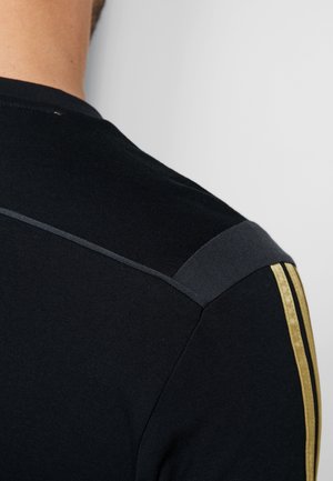 Gros plan sur le haut du dos d’une personne portant une chemise noire avec des rayures dorées sur l’épaule et une couture grise en haut.