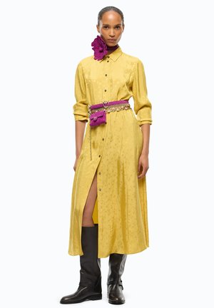 Mujer con vestido amarillo abotonado con una abertura alta, cinturón morado y accesorio de cuello, y botas negras hasta la rodilla sobre un fondo blanco.