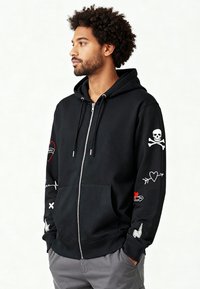 Felpa nera con zip e tasca frontale, con ricami di cuori, un teschio e frecce sulle maniche. Cappuccio con coulisse incluso.