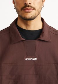Brūns teksturēts vilnas džemperis ar polo apkakli, ar melnu iekšējo slāni un izšūtu baltu Adidas logo uz krūšu.