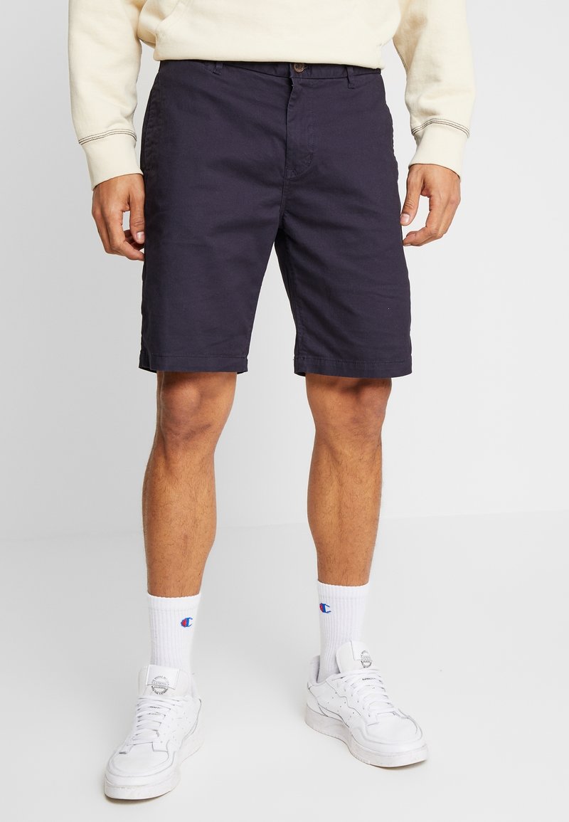 Homme portant un short bleu marine, des chaussettes blanches hautes avec un logo bleu et rouge, des baskets blanches et un sweat-shirt crème sur un fond uni.
