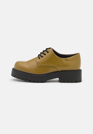 Chaussure à lacets jaune moutarde brillante avec une épaisse semelle noire à crampons, bout arrondi et petit talon sur fond clair uni.