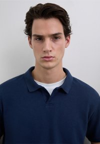 Polo azul marino con cuello clásico y mangas cortas, confeccionado en tela texturizada, superpuesto a una camiseta blanca de cuello redondo.