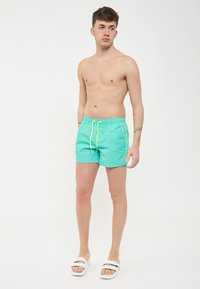 Shorts de bain turquoise avec un cordon de serrage et des accents jaunes contrastants. Le modèle se tient pieds nus, portant des sandales blanches.