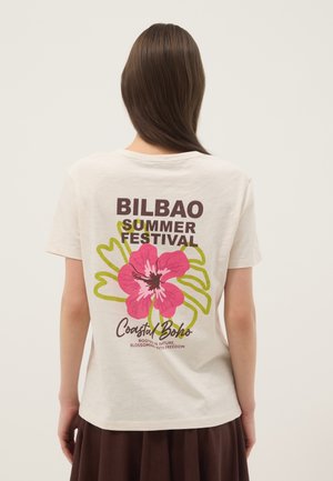 Persona con capelli lunghi castani che indossa una maglietta bianca con la scritta "Bilbao Summer Festival" e una grafica di un fiore rosa sulla schiena, abbinata a una gonna marrone.