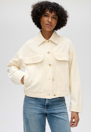 Femme aux cheveux bouclés portant une veste boutonnée en velours côtelé crème et un jean bleu, debout avec une main dans la poche devant un fond uni.
