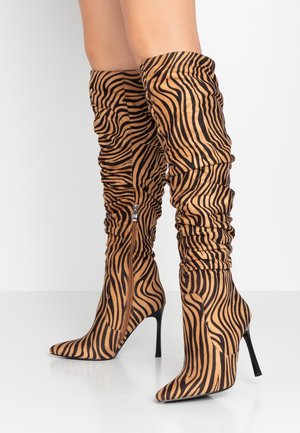 Botas holgadas hasta la rodilla, hechas de material marrón claro con rayas de tigre negras, punteras puntiagudas y tacones de aguja, con una cremallera lateral.