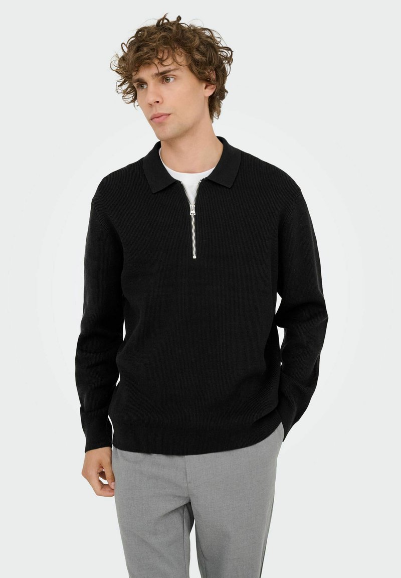 Schwarzer Strickpullover mit Reißverschlusskragen, gerippter Textur und langen Ärmeln, kombiniert mit hellgrauen Hosen.