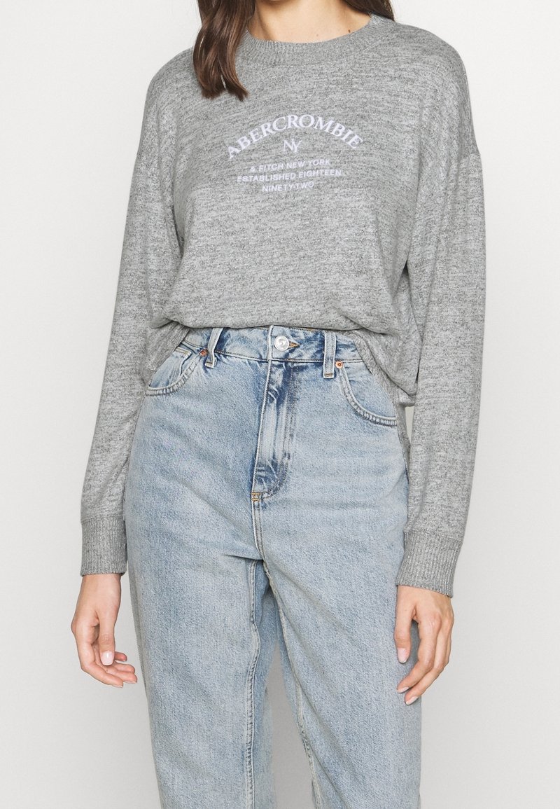 Pull en tricot gris avec un col rond et des manches longues. Présente le logo "Abercrombie" en blanc, associé à un jean bleu clair à taille haute.