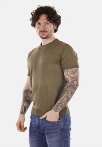 Pullover a maniche corte di colore verde oliva, con scollatura a girocollo e orlo a coste. Indossato con jeans blu, che mettono in mostra dettagliati tatuaggi sull'avambraccio.
