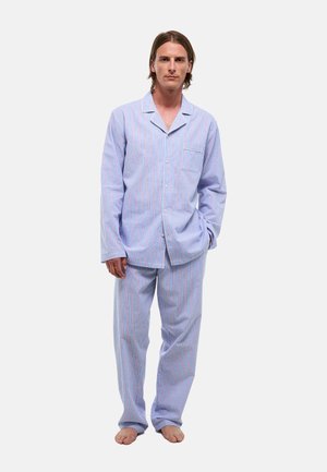 Homme debout pieds nus portant un pyjama rayé bleu clair à manches longues et pantalon, une main dans la poche, fond neutre.