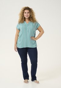 Blusa azul verdosa de botones con rayas verticales blancas, de mangas cortas, combinada con jeans azul oscuro y chanclas blancas, con un corte holgado.