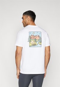 Camiseta blanca con un gráfico colorido de montañas, árboles y una tienda con el texto "TERREX" en la parte inferior, de mangas cortas y cuello redondo.