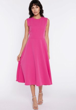 Kobieta o ciemnych włosach do ramion, ubrana w różową sukienkę midi bez rękawów i srebrne sandały na wysokim obcasie, stoi na tle białego tła.