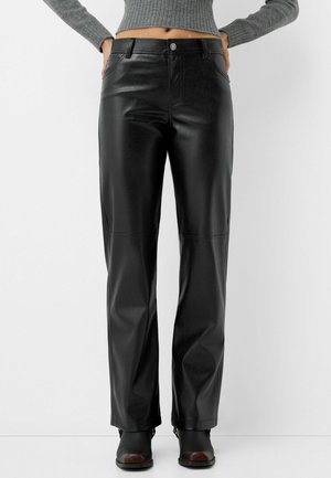 Schwarze gerade geschnittene Kunstlederhose, getragen mit schwarzen Stiefeln und einem grauen kurzen Pullover, Hände auf den Hüften ruhend.
