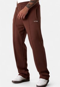 Braune Sweatpants mit glatter Textur, elastischem Bund, seitlichen Taschen und weißem Logo. Kombiniert mit weißen Sneakers mit dunkler Sohle.