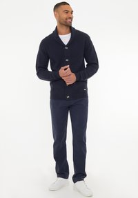 Cárdigan de punto azul marino con cuello de chal y tres botones, combinado con pantalones oscuros y zapatillas blancas. Textura suave y puños acanalados.