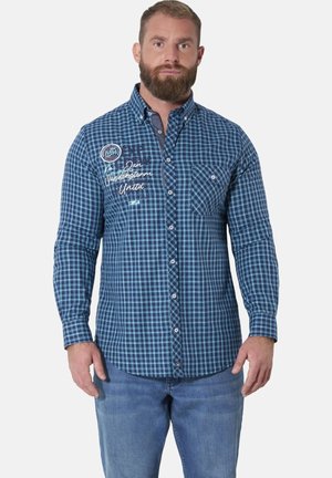 Bearded man wearing blue checkered button-up shirt with chest pocket and embroidered text, paired with blue jeans, standing facing forward.