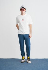 Λευκό T-shirt με στρογγυλό λογότυπο, μπλε παντελόνια φόρμας, γκρι καπέλο και πολύχρωμα γυαλιά ηλίου, σε συνδυασμό με πολύχρωμα αθλητικά παπούτσια.