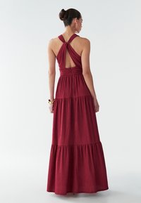 Moteris, su plaukais surištais į kuodą, vilkinti be rankovių bordo spalvos maksi suknelę su susuktais nugaros diržais ir sluoksniuotu sijonu, stovi prieš baltą foną.