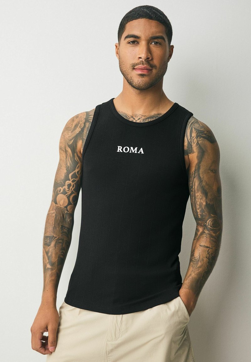 Schwarzes, geripptes Tanktop mit dem Wort "ROMA" in weißem Druck über der Brust. Das Modell hat sichtbare Armtätowierungen und helle Shorts.