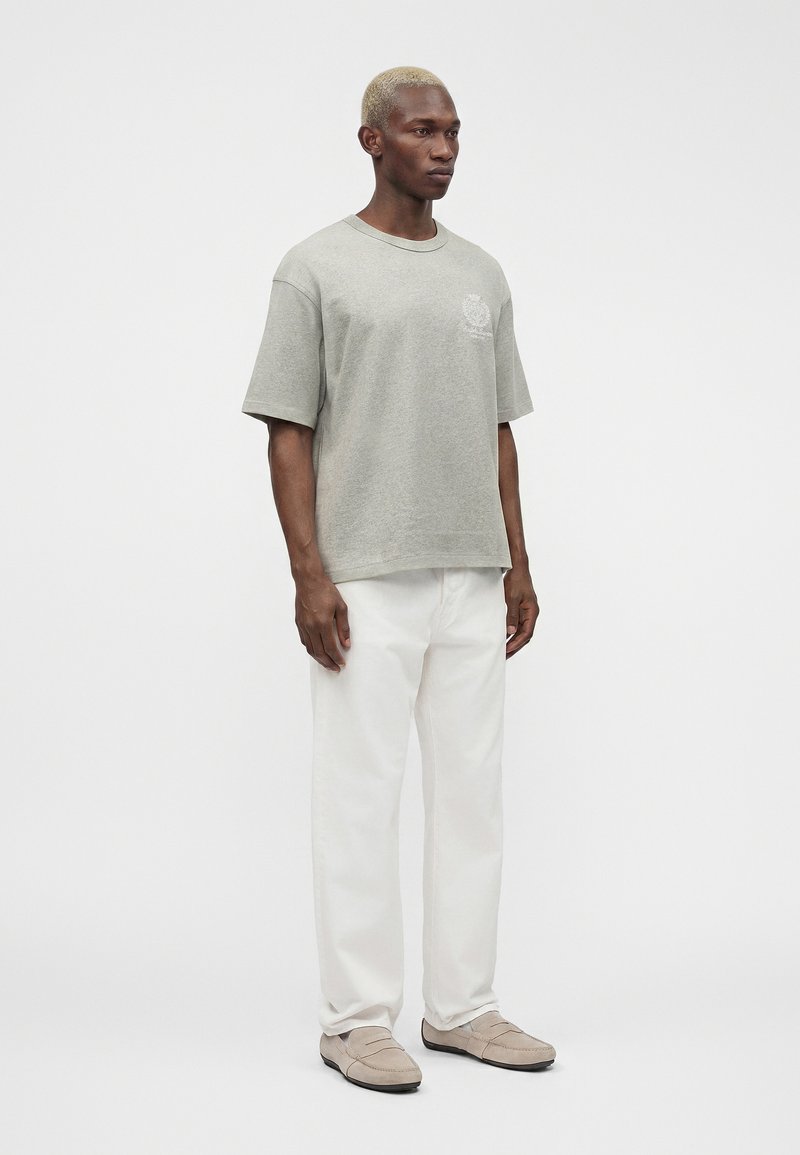 Homme aux cheveux blonds courts portant une chemise grise ample à manches courtes, un pantalon blanc et des mocassins beiges, debout devant un fond clair uni.