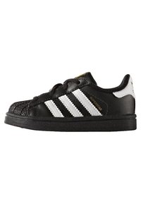 Svart Adidas Superstar-sneaker med vita ränder, skaldopp, och guld "Superstar"-text på sidan, sedd från yttersidan.