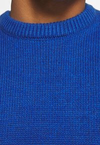 Niebieski sweter z dzianiny z okrągłym dekoltem z ribbingu. Tekstura jest gęsta i gładka, prezentująca żywy odcień niebieskiego.