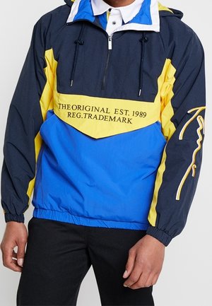 Hombre vestido con cortavientos azul, amarillo y azul marino con el texto "THE ORIGINAL EST. 1989 REG. TRADEMARK" en el frente y pantalones negros.