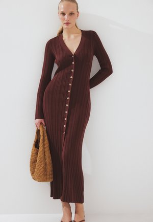 Donna che indossa un lungo vestito marrone a coste con bottoni, tiene in mano una borsa intrecciata color senape, in piedi contro un muro bianco.