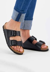Black two-strap sandals with buckles, featuring a cork footbed and textured rubber sole, paired with rolled-up denim jeans.
