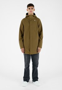 Giacca impermeabile verde oliva con colletto alto e zip frontale, indossata sopra jeans scuri e sneakers bianche; presenta un piccolo logo sul petto.