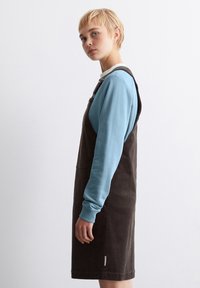 Braunes Corduroy-Latzkleid mit verstellbaren Trägern, kombiniert mit einem hellblauen Langarmshirt. Glatte Textur, knielanges Design.