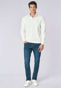 Polo a maniche lunghe, bianco, con colletto e un piccolo logo ricamato. Indossato con jeans blu e sneakers bianche.