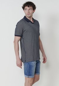 Polo tričko s námořnickou modří a bílým vzorem, s límečkem, krátkým rukávem a bočními švy. V kombinaci s ohrnutými denimovými šortkami, což vytváří ležérní outfit.