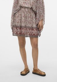 Kukkapainokuvioinen, läpikuultava mekko, jossa on kerätty vyötärö ja röyhelöhelma. Vaaleanruskeat sandaalit, joissa on ristikkäiset remmit ja musta pohja.