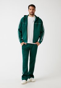 Chándal verde hecho de material texturizado, que incluye una sudadera con cremallera y pantalones con rayas laterales blancas y un patrón sutil. Camiseta blanca debajo.