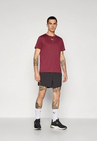 Camiseta desportiva de cor castanha com mangas curtas, combinada com calções pretos. O modelo usa sapatilhas pretas e meias brancas, exibindo tatuagens nos braços e nas pernas.