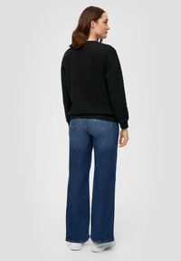 Felpa nera con scollo rotondo, abbinata a jeans di denim blu a gamba larga. Dettagli di cucitura visibili sui jeans e sneakers bianche.