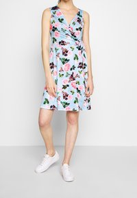 Robe portefeuille florale en bleu clair avec des roses roses et des feuilles vertes. Design sans manches, longueur genoux, stylée avec des baskets blanches.