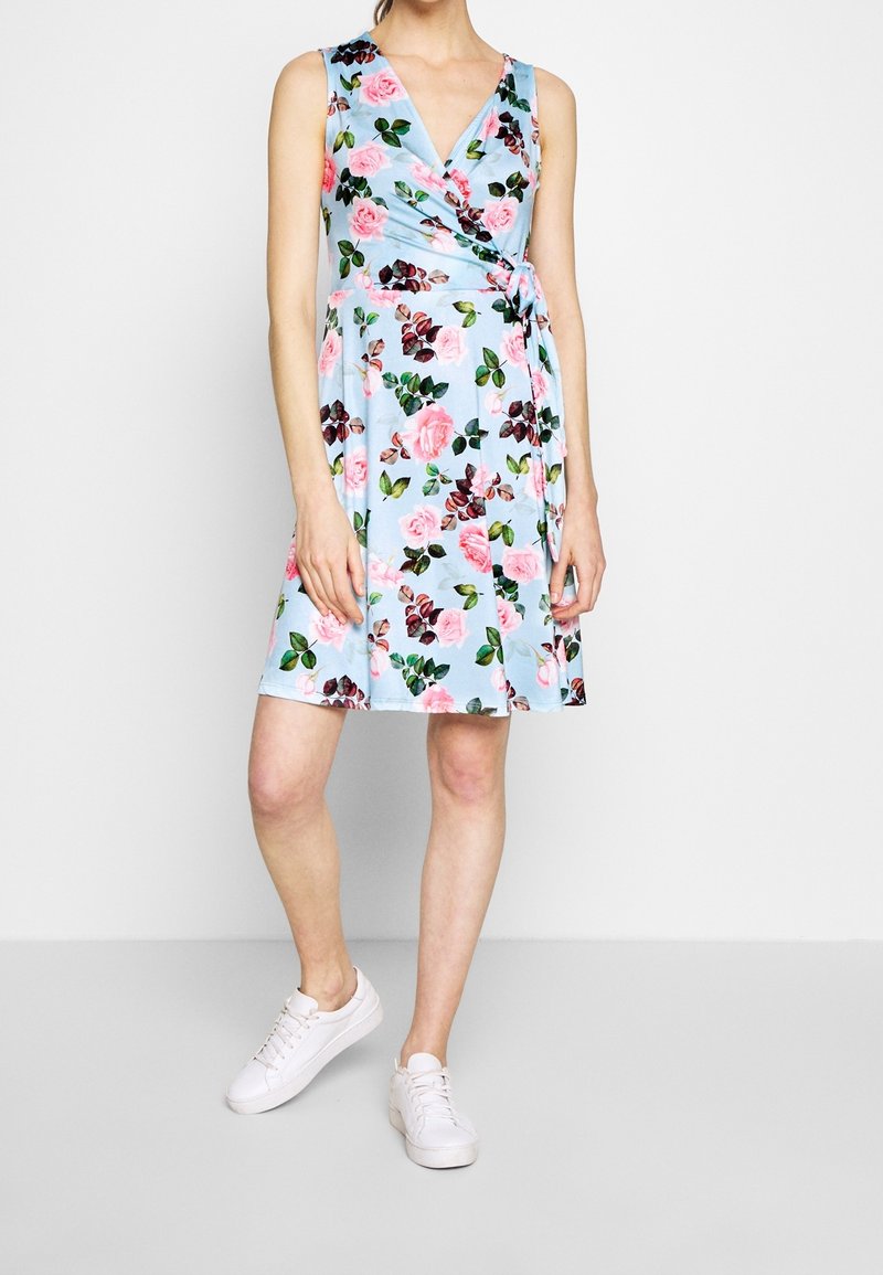 Robe portefeuille florale en bleu clair avec des roses roses et des feuilles vertes. Design sans manches, longueur genoux, stylée avec des baskets blanches.