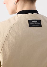 Beige jacket with a smooth texture, black collar, and a visible Ecoalf brand label on the back. Features sustainable material.