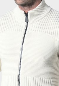 Suéter de cremallera de color beige claro con textura acanalada, cuello alto y cremallera negra. Presenta rayas verticales en la sección superior y una sección inferior lisa.