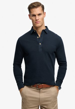 Marineblauw poloshirt met lange mouwen en een kraag met knopen, gemaakt van zachte stof. Voorzien van gouden knopen en een subtiel logo. Gecombineerd met beige broek.
