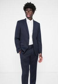 Costum bleu marin cu jachetă cu un singur rând de nasturi, rever pe notch și pantaloni. Purtați peste o cămașă albă cu guler, având o țesătură fină și un croi ajustat.