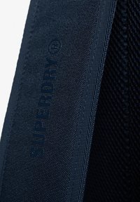 Primer plano de una correa de tela texturizada azul marino con el nombre de la marca "SUPERDRY" en relieve en vertical.