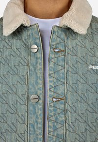 Τζάκετ τζιν με μπεζ κολάρο από shearling, μπλε ύφασμα με σχέδια ραφής σε ζικ-ζακ, ασημένια κουμπιά και λεπτομέρεια λογότυπου στο στήθος.