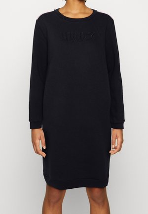 Persoon draagt een zwarte jurk met lange mouwen en een sweatshirt-stijl, met "LIU JO" reliëf op de borst, staand tegen een effen lichte achtergrond.