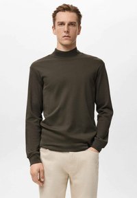 Dunkelolivgrünes, langärmeliges Rollkragenshirt aus weichem Stoff, mit lockerem Schnitt und minimalistischem Design, kombiniert mit hellbeigen Hosen.