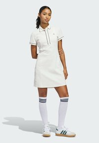 Weißes Polokleid mit schwarzem Besatz, kurzen Ärmeln und Knopfleiste; kombiniert mit weißen knielangen Socken mit marineblauen Streifen und sneakers mit grünen Akzenten.